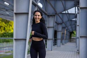 ritratto di un' giovane attivo donna, latino americano donna con Riccio capelli nel un' tuta da ginnastica sorridente e guardare a il telecamera, sportiva in esecuzione nel il parco vicino il urbano costruzione. foto