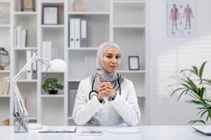 un' professionale ritratto di un' fiducioso femmina medico indossare un' hijab, seduta nel sua ben organizzato clinica ufficio, illustrare inclusività e diversità nel assistenza sanitaria. foto