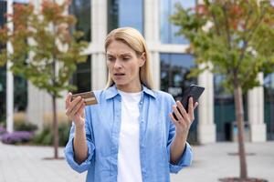 ingannato donna al di fuori ufficio edificio negato i soldi trasferimento, maturo femmina lavoratore con Telefono e banca credito addebito carta in piedi, irritato e infelice bionda. foto