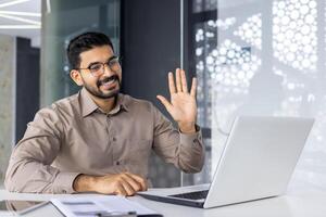 un' allegro maschio professionale nel un' inteligente camicia onde calorosamente a il suo il computer portatile durante un' virtuale incontro nel un' moderno ufficio ambientazione. foto