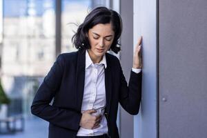 giovane donna d'affari sofferenza a partire dal stomaco dolore, in piedi vicino il ufficio costruzione, pendente contro il parete, Tenere sua corpo. foto