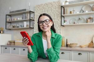 giovane bellissimo donna nel bicchieri e verde camicia nel il cucina utilizzando un' rosso Telefono foto