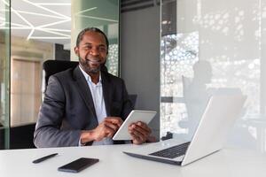 ritratto di riuscito africano americano uomo d'affari dentro ufficio, capo con tavoletta computer sorridente e guardare a telecamera, uomo a posto di lavoro contento con finanziario risultati e conquiste. foto