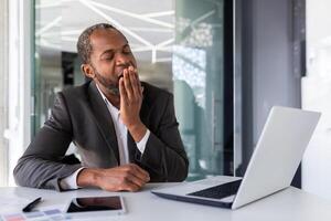 stanco uomo d'affari manca noioso opera e ogni giorno, africano americano maturo esperto uomo nel attività commerciale completo da uomo sbadiglia, a posto di lavoro dentro ufficio. foto