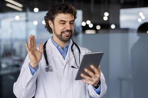 sorridente maschio medico nel un' laboratorio cappotto con stetoscopio utilizzando un' tavoletta per un' virtuale saluto nel un' moderno clinica ambientazione. foto