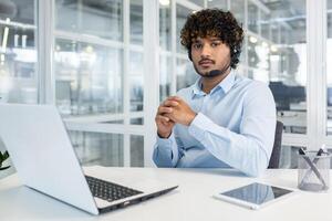 fiducioso maschio cliente supporto rappresentante con Riccio capelli, utilizzando un' cuffia, lavori su un' il computer portatile nel un' moderno ufficio ambientazione. foto