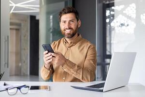 ritratto di maturo riuscito uomo d'affari dentro ufficio, maschio guardare a telecamera sorridente, lavoratore nel camicia Tenere Telefono, utilizzando in linea applicazione. foto