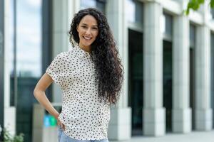 sorridente giovane ispanico donna con Riccio capelli godendo sua pranzo rompere al di fuori un ufficio. fiducioso e professionale. foto