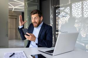 scioccato e contento giovane uomo d'affari uomo seduta nel il ufficio a il scrivania e guardare sorpreso a il Telefono schermo, emotivamente contento con il notizia e successo. foto