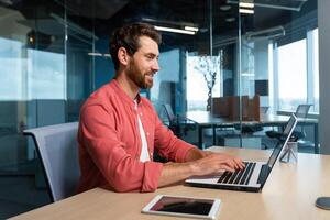 riuscito uomo d'affari nel rosso camicia felicemente Lavorando con il computer portatile dentro ufficio, maturo uomo con barba a posto di lavoro digitando su tastiera sorridente soddisfatto con opera risultati e risultato. foto