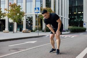 un' giovane asiatico uomo corre nel il mattina attraverso il strade di il città, sta e detiene il suo gamba, si sente acuto dolore e spasmo nel il suo ginocchio, ha un' gli sport lesione. foto