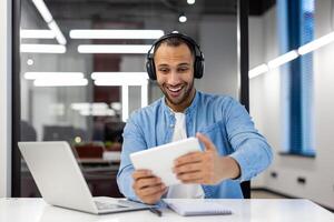 sorridente giovane latino americano uomo seduta a un' scrivania nel il ufficio indossare cuffie e con entusiasmo giocando in linea Giochi su un' tavoletta. foto