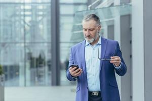 anziano bello dai capelli grigi uomo nel un' completo da uomo Tenere un' Telefono e occhiali. concentrato legge il notizia su il Telefono, sembra a messaggi. esso è collocato vicino un' moderno ufficio centro. foto