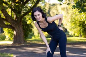 irritato giovane donna in piedi nel il parco piegato al di sopra di e Tenere sua mano dietro a sua Indietro, sofferenza a partire dal acuto dolore e distorsione dopo jogging e Lavorando fuori. foto