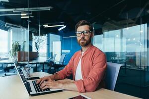 ritratto di un' riuscito programmatore dentro il ufficio, un' uomo con un' barba, bicchieri e un' rosso camicia sembra a il telecamera, un' sviluppatore ingegnere usi un' il computer portatile per Scrivi codice nel un' programmazione struttura. foto