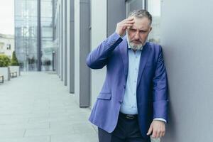 anziano uomo, lavoratore, uomo d'affari nel un' completo da uomo sta vicino il ufficio centro, Leans contro il parete, detiene il suo mano dietro a il suo testa, si sente un' male alla testa. nervi, fatica a opera. foto