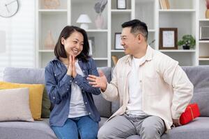 contento asiatico famiglia uomo e donna insieme a casa seduta su divano nel vivente camera, marito dando regalo per moglie nel celebrazione, coppia abbracciare e contento. foto