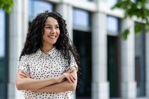 un' professionale donna nel un' polka punto camicia, con fiducia in piedi con attraversato braccia al di fuori un' contemporaneo ufficio costruzione. foto