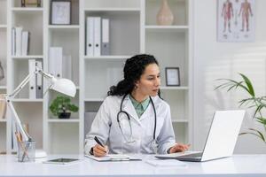 professionale ispanico femmina medico nel un' bianca cappotto scrittura mentre Lavorando su un' il computer portatile nel sua ufficio. foto