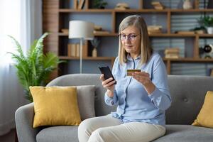maturo donna seduta su divano a casa, utilizzando smartphone e Tenere credito carta per in linea acquisti. foto