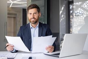 fiducioso, contento maschio contabile rivedere lavoro d'ufficio con un' Sorridi nel un' luminosa, contemporaneo ufficio ambientazione. foto