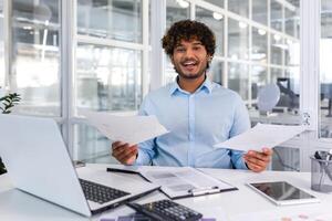 soddisfatto con il risultato finanziere uomo d'affari detiene nel il suo mani e esamina contratti e rapporti sorridente e felice, uomo nel un' casuale camicia lavori dentro il ufficio utilizzando un' il computer portatile nel scartoffie. foto