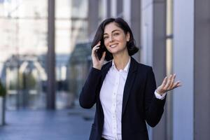 un' giovane donna d'affari è parlando su il strada vicino un ufficio edificio su un' mobile Telefono, discutere, Esprimere a gesti con sua mani. foto