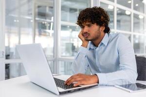 irritato pensiero uomo d'affari Lavorando con il computer portatile dentro moderno ufficio, uomo frustrato soluzione difficile decisione a posto di lavoro, ispanico uomo con Riccio capelli grave nel camicia. foto