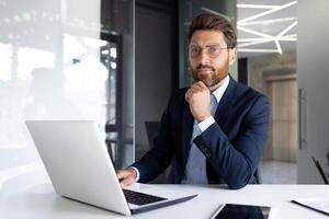 ritratto di un' grave giovane attività commerciale uomo, un' avvocato seduta nel il ufficio a un' Lavorando tavolo con un' il computer portatile e con fiducia guardare a il telecamera. foto