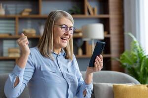 entusiasta femmina godendo successo con pollici su mentre Tenere un' mobile Telefono nel un' accogliente vivente camera. foto