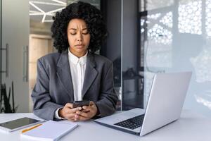 irritato giovane africano americano donna Lavorando nel il ufficio su un' computer portatile, seduta a il scrivania, Tenere il Telefono e guardare purtroppo a il schermo. foto