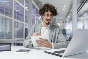 professionale giovane maschio con Riccio capelli Lavorando a il computer portatile nel un' aziendale ufficio ambientazione, mostrando messa a fuoco e competenza. foto