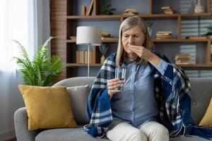 triste anziano signora seduta su imbottito divano sotto coperta e ottenere pronto per assunzione medicina con bicchiere di acqua. lottando donna sofferenza a partire dal mal di testa e utilizzando antidolorifici durante malato partire. foto