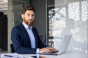 ritratto di un' grave e sicuro di sé giovane uomo d'affari seduta nel il ufficio a il tavolo, digitando su un' computer portatile, Lavorando con clienti e documenti, guardare a il telecamera. foto