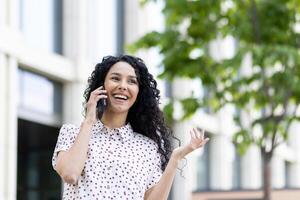 giovane bellissimo donna parlando su il Telefono avvicinamento mentre a piedi nel il città, attività commerciale donna sorridente con soddisfazione, riuscito latino americano donna con Riccio capelli su un' soleggiato giorno. foto