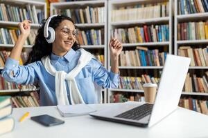 eccitato giovane donna con cuffie danza con gioia a un' biblioteca scrivania, circondato di libri. lei trasuda positività e un' amore per musica mentre Lavorando. foto