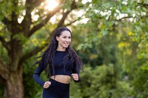 giovane motivata e gioioso donna jogging nel il mattina nel un' pubblico parco tra alberi, ispanico donna con Riccio capelli, sottile nel un' tuta. foto