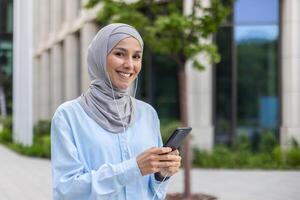 sorridente giovane musulmano femmina professionale nel hijab coinvolgente con smartphone nel davanti di un ufficio edificio nel il città. foto