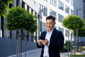 asiatico giovane maschio uomo d'affari in piedi su il strada vicino il ufficio centro nel un' attività commerciale completo da uomo e utilizzando un' mobile Telefono, sorridente e digitando e guardare a il schermo. foto