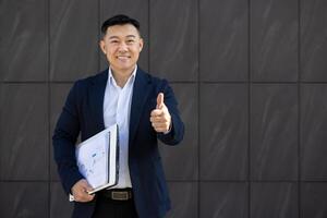 sorridente asiatico uomo d'affari nel completo da uomo dando un' pollici su cartello mentre Tenere un' documento con grafici, in piedi contro un' grigio sfondo. foto