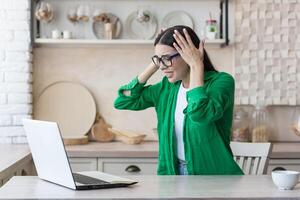 stressato donna a casa Lavorando nel cucina utilizzando computer portatile, avuto cattivo notizia in linea foto