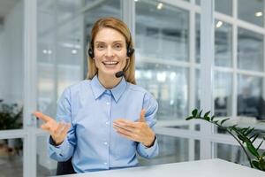 sorridente professionale femmina con cuffia fabbricazione gesti su un' chiamata nel un' luminosa ufficio ambiente, guardare a telecamera. foto
