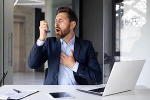 un' giovane uomo d'affari soffre a partire dal asma e allergie. seduta nel il ufficio a il scrivania e utilizzando un inalatore. foto