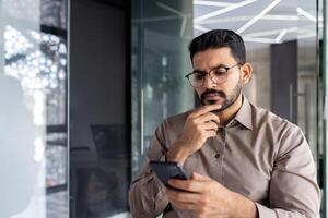 uomo d'affari pensiero mentre seduta dentro ufficio a tavolo, uomo è concentrandosi e sul serio lettura in linea utilizzando un applicazione su Telefono, il giovane capo è Tenere un' smartphone nel il suo mani. foto