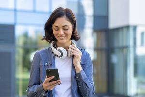 un' giovane femmina alunno con un' allegro espressione usi sua smartphone e indossa cuffia, in piedi all'aperto vicino un' moderno costruzione. concetto di tecnologia nel quotidiano vita. foto