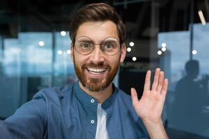 ritratto di riuscito uomo d'affari nel casuale camicia e bicchieri uomo guardare a smartphone telecamera e sorridente, telecamera Visualizza chiamata parlando con amici dentro ufficio a distanza. foto