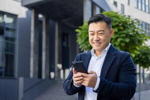 positivo asiatico uomo d'affari nel un' completo da uomo godendo un' rompere all'aperto con il suo mobile Telefono, trasudante fiducia e successo. foto