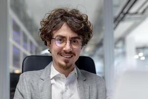 sorridente giovane uomo d'affari con Riccio capelli e bicchieri Lavorando a il suo computer nel un' contemporaneo ufficio ambientazione. foto