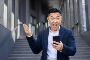 felicissimo asiatico uomo d'affari nel completo da uomo festeggiare bene notizia su il suo Telefono a un urbano scala ambientazione. foto