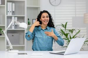 gioioso donna d'affari con Riccio capelli nel un' blu camicia si siede a sua ufficio scrivania, Esprimere a gesti un' 'chiamata Me' cartello e sorridente energeticamente. foto